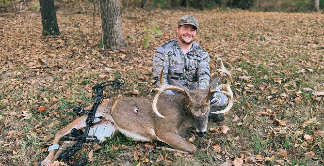 A hunter in camo sits on a forest floor beside a large deer with antlers, bow on the ground. The scene conveys a successful hunt in autumn.