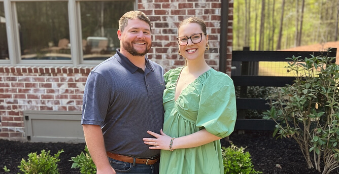 A couple stands together outside in front of a brick building, smiling at the camera. The woman is wearing a green dress with puffed sleeves, and the man is in a blue polo shirt. There are plants around them and trees in the background.