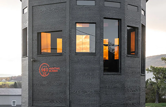 A black, octagonal building with multiple windows reflects a vibrant sunset. Text on the exterior reads "468 Printing House." Trees and hills are visible nearby.