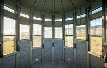 Interior of a round, windowed room with a panoramic view of a sunny landscape. Tall windows and a curved ceiling create a spacious ambiance.
