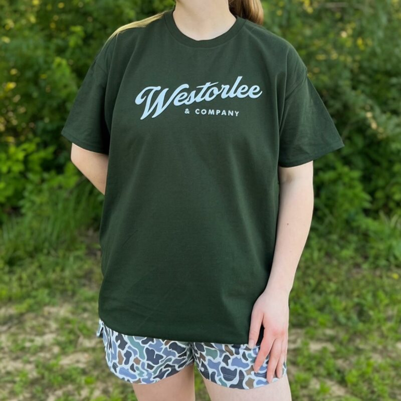 Person wearing a dark green T-shirt with "Westerlee & Company" text, paired with camo shorts, standing outdoors against a leafy background.
