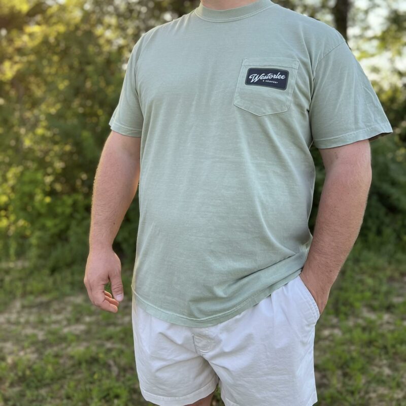 A person wearing a light green T-shirt with a logo on the pocket and white shorts is standing outdoors. Sunlight filters through trees in the background.