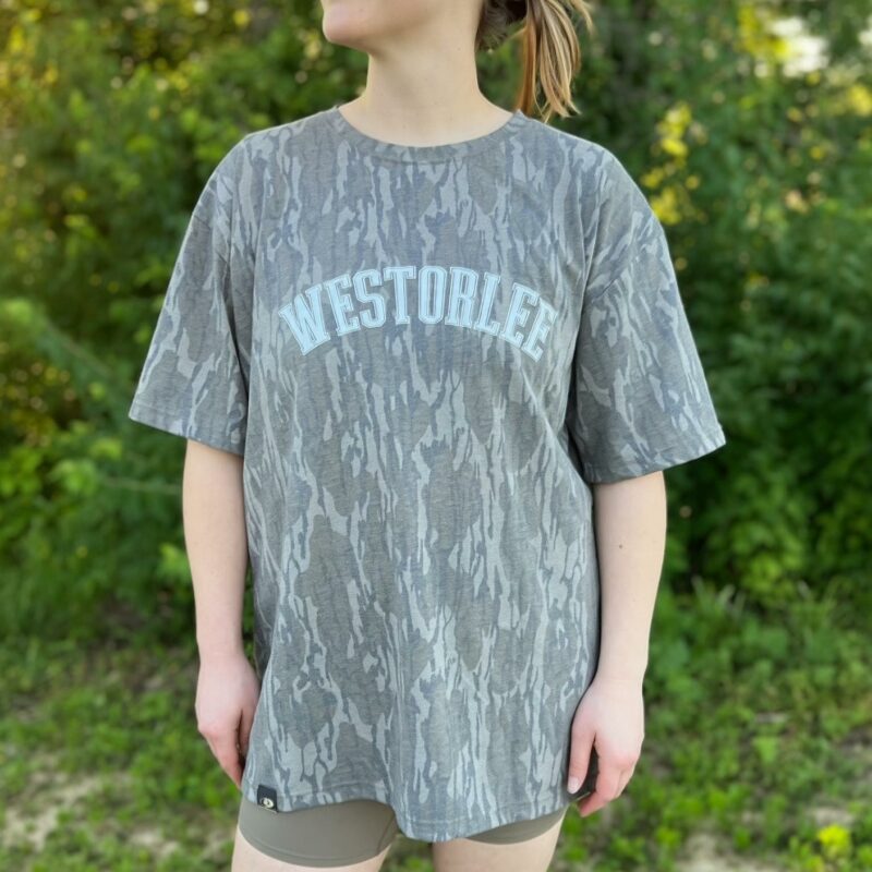 A person stands outdoors wearing a gray camouflage-patterned shirt with "WESTORLEE" in white text. The background is lush green foliage.