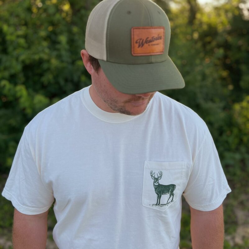 Man in a white T-shirt with a deer graphic on the pocket, wearing a green cap outdoors. Background features lush green foliage in soft focus.